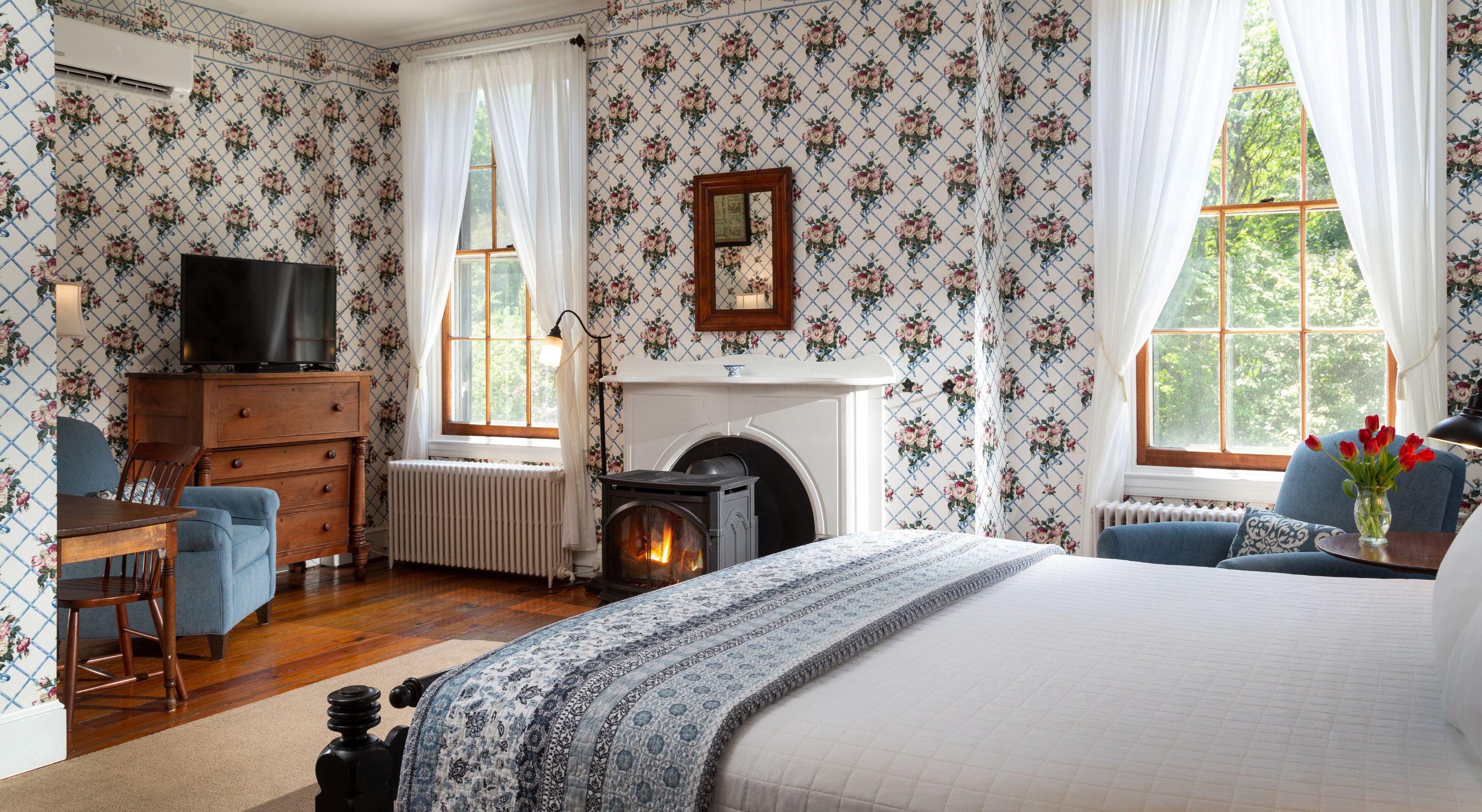 Spacious guest room with floral wallpaper, natural light, fire stove, table for two, TV and chair, and comfy bed