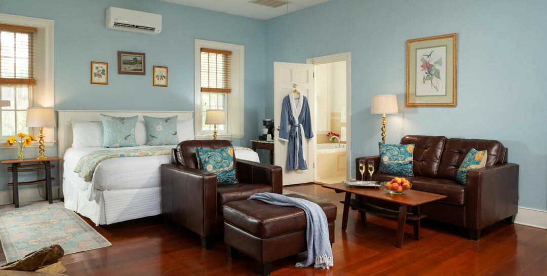 Light blue Sunrise Room with off-white king bed, windows with blinds, wood floors, and leather sitting area.